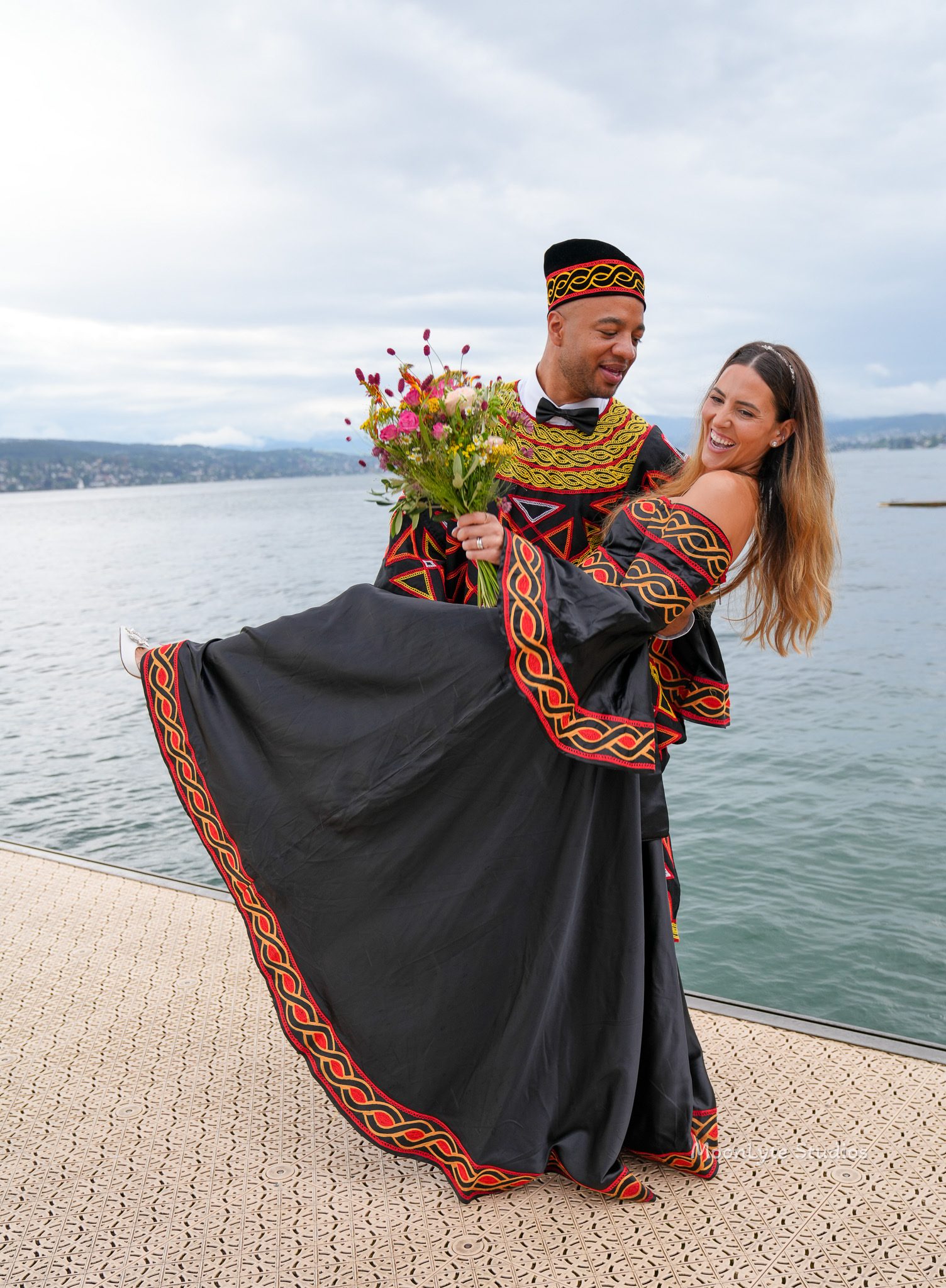 Joyful couple in black traditional attire with red and gold embroidery sharing a playful dip by the water